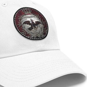 May include: White baseball cap with a round embroidered patch featuring a red, white, and black eagle with a crown on top. The eagle is surrounded by a wreath of green leaves.