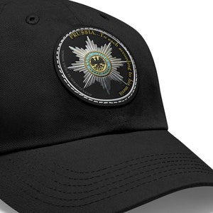 May include: Black baseball cap with a circular patch featuring a silver star with a blue and gold eagle design. The patch has the text "PRUSSIA... To seek and to find the way from the heart" around the star.
