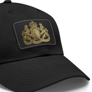 Puede incluir: Una gorra de béisbol negra con un escudo bordado en oro que presenta una corona y leones. El escudo está rodeado por un borde blanco cosido.