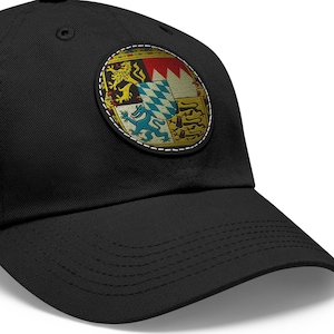 Puede incluir: Una gorra de béisbol negra con un parche redondo cosido que presenta el escudo de armas de Baviera. El parche es amarillo, rojo, azul y blanco.
