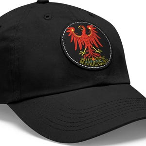 May include: Black baseball cap featuring a circular patch with a red eagle emblem. The patch includes the word "BRANDENBURG" in gold lettering. The cap has a curved brim and a button on top.