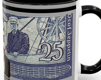 May include: A white ceramic mug with a black interior and handle. The mug displays a vintage blue and grey graphic of a man, a zeppelin, and the number 25. The text "Graf Zeppelin" is printed on the side.