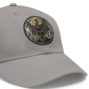 May include: A gray baseball cap with a black and gold embroidered patch featuring a stylized eagle with a crown and a shield.