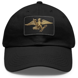 May include: Black baseball cap featuring a gold-colored eagle emblem on a rectangular patch. The eagle is depicted with outstretched wings, holding arrows. The cap has a curved brim and a classic, adjustable design.