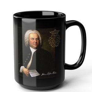 May include: Black ceramic mug featuring a portrait of Johann Sebastian Bach, a German composer, on the front. The mug has a white handle and a gold crown with a musical symbol above the portrait.