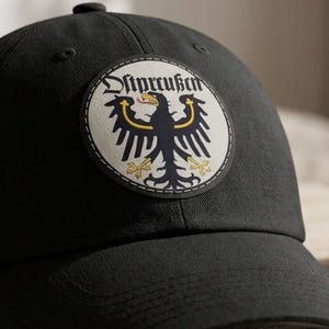 May include: A black baseball cap featuring a circular patch on the front. The patch displays a black eagle with yellow accents on a cream background, with the text Ostpreußen written in black gothic script above the eagle's head.