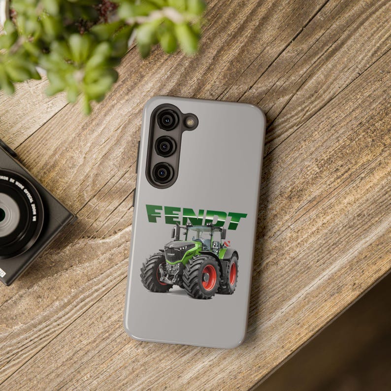 May include: A gray iPhone 13 Pro Max phone case features a green Fendt tractor graphic. The case showcases the Fendt logo in green text above a detailed image of a modern green and black tractor with large tires. The phone case is designed for an iPhone 13 Pro Max, with a camera cutout visible. The overall design is sleek and simple, ideal for fans of agricultural machinery and the Fendt brand.