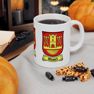 May include: White ceramic coffee mug with a red and yellow coat of arms featuring a tower and the text "Memml". The mug is sitting on a white surface with orange and black candy.