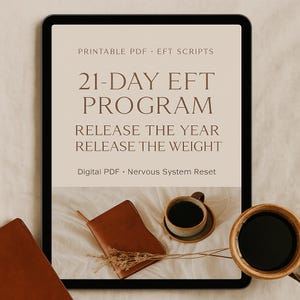 May include: A digital tablet displays a beige graphic with the text "21-DAY EFT PROGRAM" and "RELEASE THE YEAR RELEASE THE WEIGHT." A brown leather journal, a cup of coffee, and a wooden mug with coffee are arranged on a cream-colored surface.