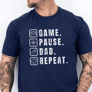 May include: Dark blue t-shirt featuring white text and icons. The text says "GAME. PAUSE. DAD. REPEAT." with icons representing a game controller, pause button, baby bottle, and a recycling symbol. The shirt has short sleeves.