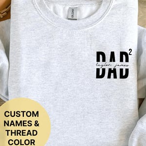 Op de afbeelding: Grijs sweatshirt met de tekst "DAD²" en "Taylor James" in een kleiner lettertype eronder. De tekst is zwart. Het sweatshirt is een ronde hals stijl.