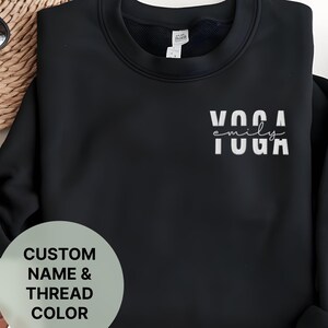Puede incluir: Sudadera negra con el texto "emily yoga" en letras blancas. La sudadera es de cuello redondo y tiene un corte holgado.