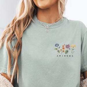 Op de afbeelding: Lichtgroen t-shirt met een bloemenontwerp en de letters A, J, N en S. Het shirt heeft een ronde hals en korte mouwen. Het bloemenontwerp bevat blauwe, paarse, roze en gele bloemen.