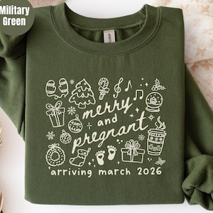 Puede incluir: Una sudadera de cuello redondo color verde militar con un estampado gráfico en color crema de iconos navideños que incluyen un árbol de Navidad, regalos y una taza de café. El texto dice "merry and pregnant" y "arriving march 2026".