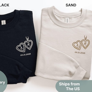 Puede incluir: Se muestran dos sudaderas, una negra y otra color arena. Cada una presenta un diseño bordado de dos medallones en forma de corazón conectados con las iniciales "A" y "M" y la fecha "09.21.2025". La palabra "Bordado" es visible a la izquierda.