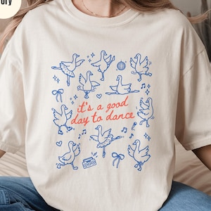 May include: Ivory-colored t-shirt with a blue graphic of dancing geese and the phrase "it's a good day to dance" in red. The design includes musical notes, bows, and a disco ball. The shirt is a casual, comfortable style.