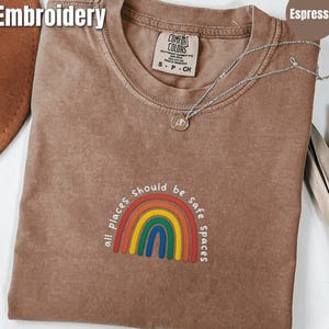 Puede incluir: Camiseta color espresso con el texto "all places should be safe spaces" bordado sobre un diseño de arcoíris. La camiseta tiene una etiqueta Comfort Colors y un collar plateado. La palabra "Embroidery" está en la esquina superior izquierda.