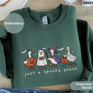 Puede incluir: Sudadera verde bosque con bordado de gansos de Halloween. El diseño presenta gansos disfrazados de calabaza, fantasma, bruja y momia, con el texto "just a spooky goose". La sudadera es de material suave.