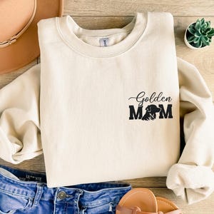 Puede incluir: Una sudadera de color crema con el texto "Golden Mom" y una silueta de un perro golden retriever.