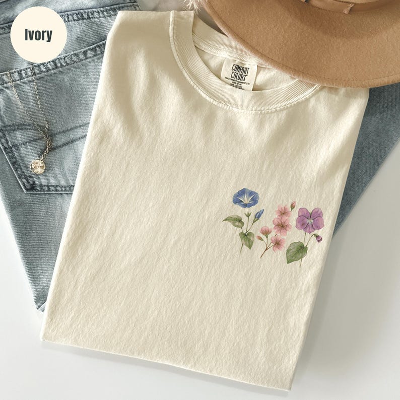 Op de afbeelding: Ivoor kleurig T-shirt met een bloemenontwerp met blauwe, roze en paarse bloemen. Het shirt is opgevouwen op een blauwe jeans. Een bruine hoed en een zilveren ketting zijn ook zichtbaar.
