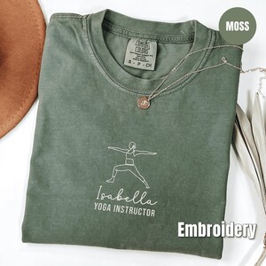 Puede incluir: Una camiseta Comfort Colors verde musgo con un diseño de pose de yoga bordado en blanco y el nombre "Isabella, Instructora de Yoga". Un collar plateado con un colgante descansa sobre la camisa. La palabra "Bordado" está en la esquina inferior derecha.