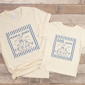 May include: Two cream-colored t-shirts. One reads "mama goose" with a blue graphic of geese and the text "Mommy & Liam". The other reads "little goose" with a similar design. Both shirts have the year "2026".