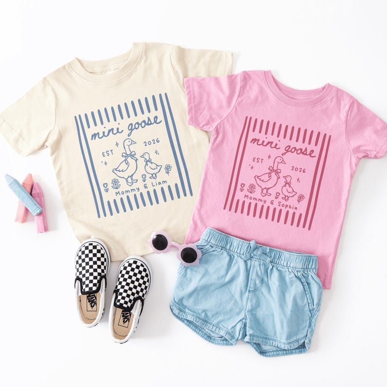 May include: Two children's t-shirts, one cream and one pink, with a "mini goose" graphic. The cream shirt has a blue design, and the pink shirt has a red design. Also pictured are checkered sneakers, sunglasses, and denim shorts.