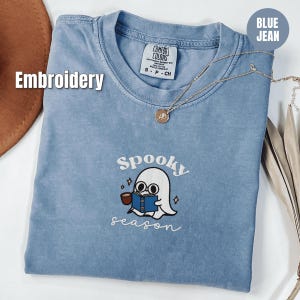 Puede incluir: Una camiseta azul vaquera Comfort Colors con un diseño bordado. El diseño presenta un fantasma de dibujos animados leyendo un libro con las palabras "spooky season" debajo. Un collar dorado está drapeado sobre la camisa.