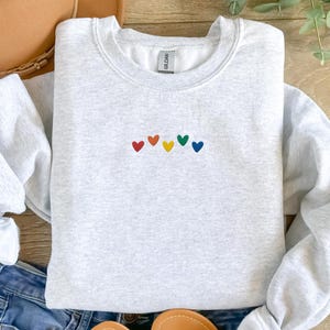 Puede incluir: Una sudadera gris con un arcoíris de pequeños corazones bordados en la parte delantera.