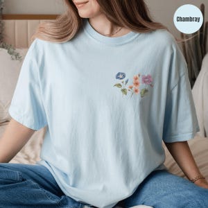 Op de afbeelding: Lichtblauw T-shirt met een klein bloemenontwerp op de borst. Het ontwerp toont bloemen in aquarel-stijl in blauwe, roze en paarse tinten. Het shirt is gemaakt van een zacht materiaal en heeft korte mouwen. Het woord "Chambray" staat in een cirkel rechtsboven.
