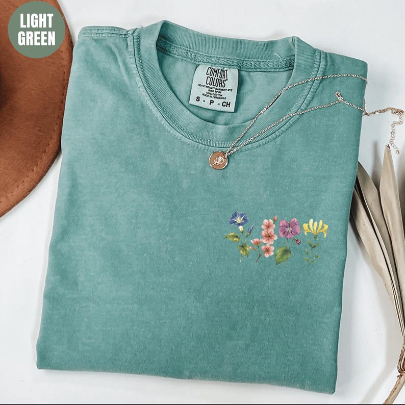 Op de afbeelding: Lichtgroen t-shirt met een bloemenontwerp en een Comfort Colors label. Het shirt heeft een kleine bloemenarrangement in roze, paars en geel. Een zilveren ketting met een hanger is over het shirt gedrapeerd.