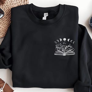 Puede incluir: Sudadera negra con un gráfico blanco de un libro abierto con flores y un diseño de luna creciente. El texto "Read" está impreso en el libro.