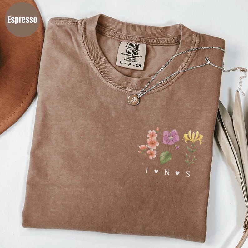 Op de afbeelding: Espresso-kleurig T-shirt met een bloemenontwerp en de initialen JNS. Het shirt heeft een ronde hals en korte mouwen. Een zilveren ketting met een kleine hanger is over het shirt gedrapeerd. Het shirt is gemaakt van een zacht, comfortabel materiaal.