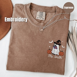 Puede incluir: Camiseta color espresso con un diseño bordado de fantasma de Halloween. El fantasma lleva un sombrero de bruja y lee un libro, flanqueado por dos calabazas. El texto "Mrs. Smith" está debajo. Un collar plateado con un colgante descansa sobre la camiseta.