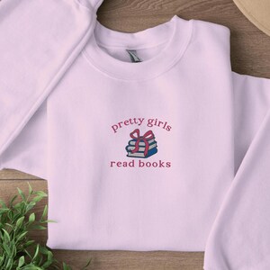 Puede incluir: Una sudadera rosa claro con el texto "pretty girls read books" y una pila de libros con una cinta rosa en la parte superior.