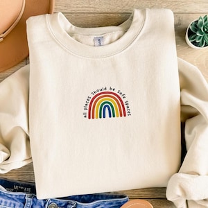Puede incluir: Una sudadera blanca con un gráfico de arcoíris y el texto "Todos los lugares deberían ser espacios seguros".