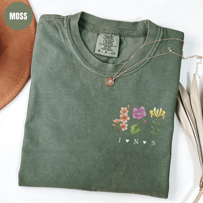Op de afbeelding: Een mosgroen t-shirt met een bloemenontwerp en de initialen J, N en S. Het shirt heeft een ronde hals en korte mouwen. Een gouden ketting met een hanger rust op het shirt. Het shirt is gemaakt van een zacht materiaal.