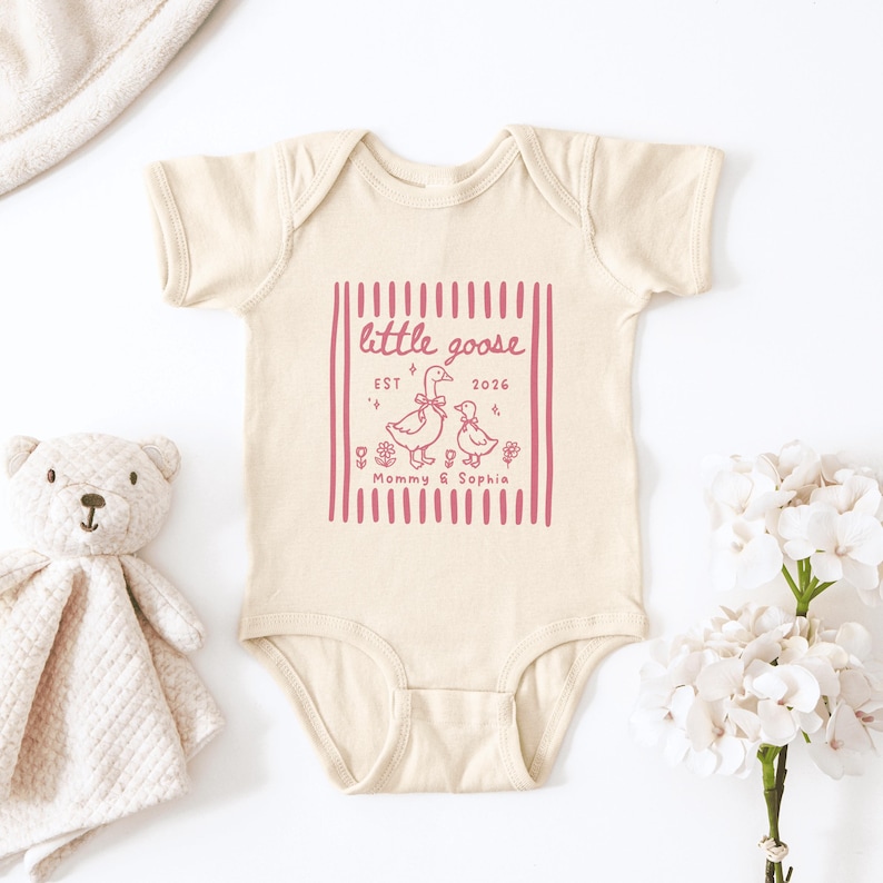 May include: Cream-colored baby onesie with a pink graphic that reads "little goose" and "Mommy & Sophia." The design includes two geese, flowers, and the year 2026. A teddy bear and flowers are in the background.