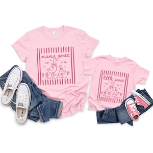 May include: Pink t-shirts with matching designs, one adult and one child size. The adult shirt says "mama goose" with a goose graphic, and the child's shirt says "little goose." Both shirts are paired with blue jeans and sneakers.