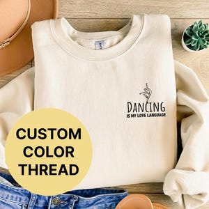 May include: A cream-colored sweatshirt with the text "Dancing is my love language" and a silhouette of a dancer. The sweatshirt has a custom color thread option.