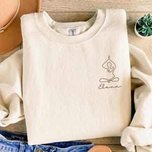 Puede incluir: Una sudadera de color crema con un dibujo lineal marrón de una persona en una postura de yoga y el nombre "Elena" escrito debajo.