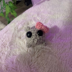 May include: A small grey crocheted animal with black eyes and a pink crocheted bow on its head. The animal is sitting on a white fluffy surface.