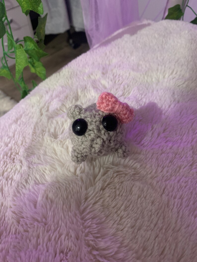 May include: A small grey crocheted animal with black eyes and a pink crocheted bow on its head. The animal is sitting on a white fluffy surface.