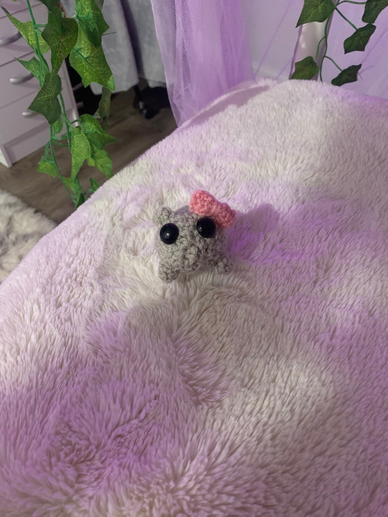 May include: A small grey crocheted plush toy with black eyes and a pink heart on its head. The toy is sitting on a white fluffy surface.