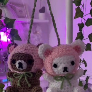 Op de afbeelding: Twee gehaakte pluche speelgoedjes die aan een groene touwtje hangen. Het speelgoedje links is bruin met een roze kap en een groene strik. Het speelgoedje rechts is wit met een roze kap en een groene strik.