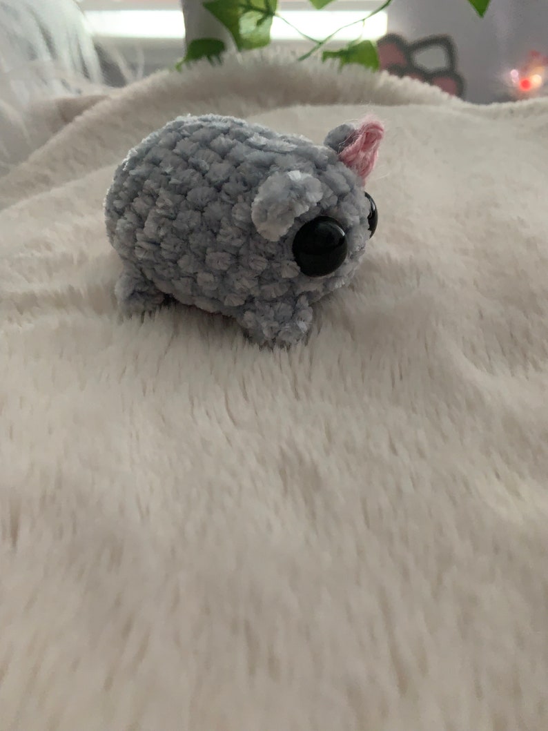 May include: A small, grey, crocheted plush toy with large black eyes and a pink ear. The toy is sitting on a white, fluffy surface.