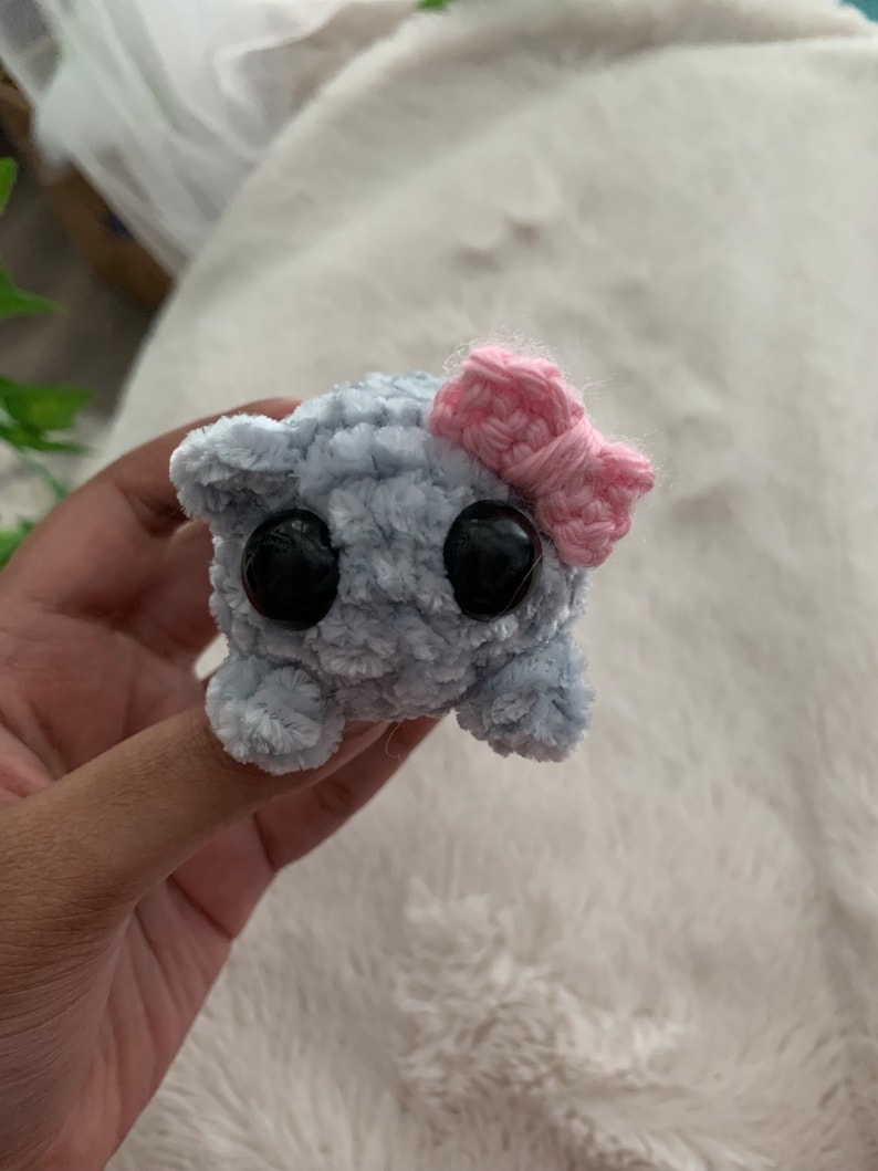May include: A small, grey, fuzzy, crocheted toy with large black eyes and a pink crocheted bow on its head.