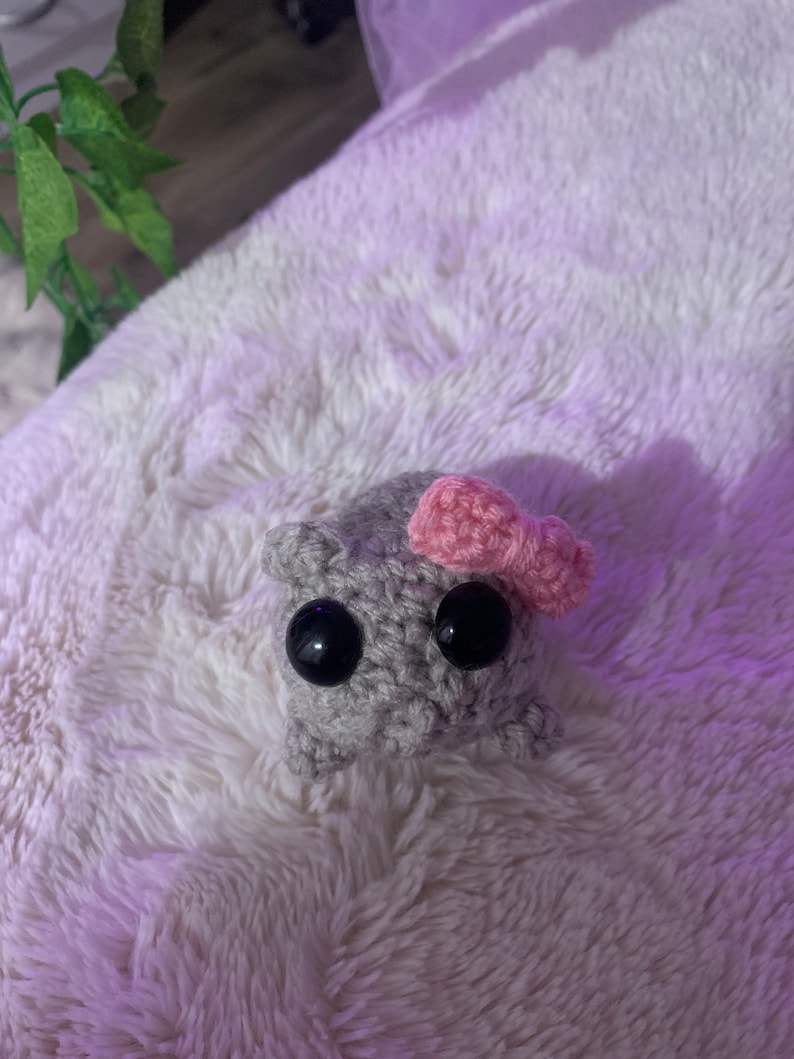 May include: A grey crocheted stuffed animal with large black eyes and a pink bow on its head.
