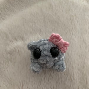 May include: A small grey crocheted plush toy with large black eyes and a pink crocheted bow on top.