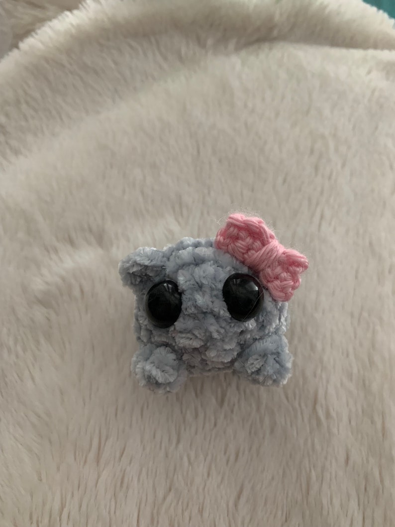 May include: A small grey crocheted plush toy with large black eyes and a pink crocheted bow on top.